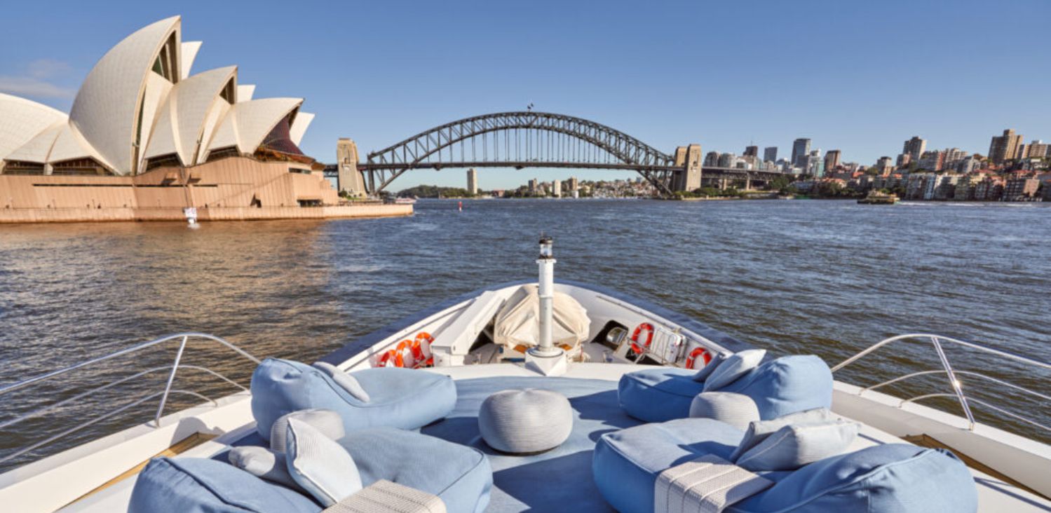 View from the bow of a boat showing cushioned seats and a railing, sailing towards the sydney opera house and harbour bridge under a clear blue sky.